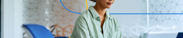 The image shows a professional woman sitting at a desk and working on a laptop in a modern office setting.