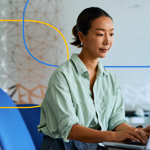 The image shows a professional woman sitting at a desk and working on a laptop in a modern office setting.