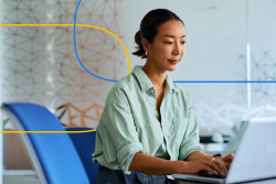 The image shows a professional woman sitting at a desk and working on a laptop in a modern office setting.