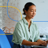 The image shows a professional woman sitting at a desk and working on a laptop in a modern office setting.
