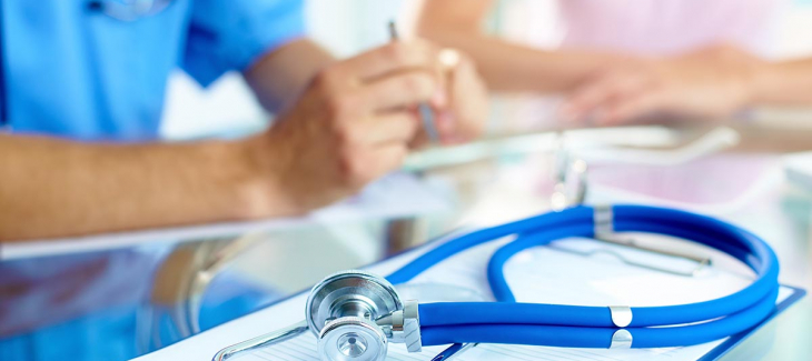 Close-up of stethoscope on a clipboard with a healthcare professional in blue scrubs discussing paperwork with a patient in the background