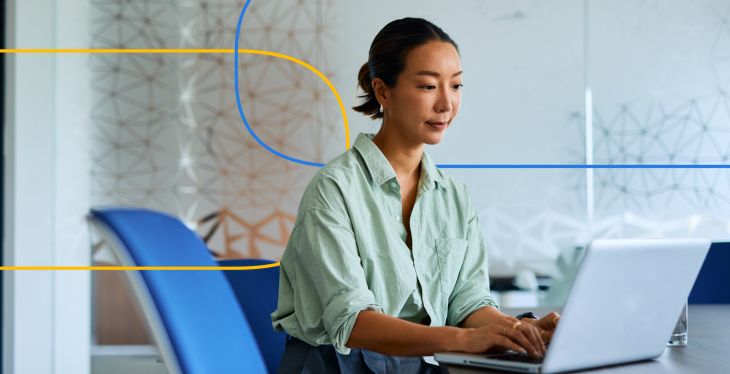 The image shows a professional woman sitting at a desk and working on a laptop in a modern office setting.